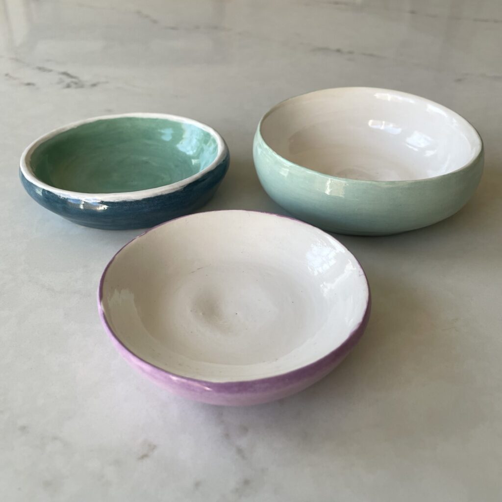 Three small homemade bowls. The top left bowl has a dark teal outside, lighter teal inside, and an uneven white rim on the top. The top right bowl has a light teal outside and white inside. The bottom bowl has a light purple outside and white inside.